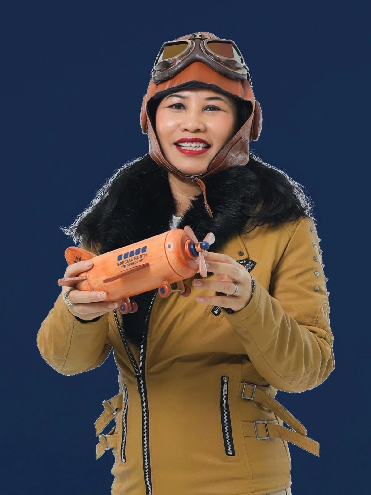 Aida Ismail in a leather flying helmet and goggles and a shearling flight jacket, holding a wooden model aeroplane