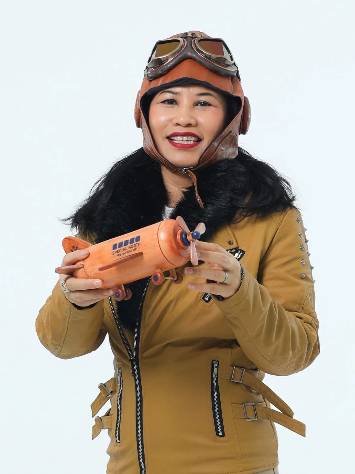 Aida Ismail in a leather flying helmet and goggles and a shearling flight jacket, holding a wooden model aeroplane